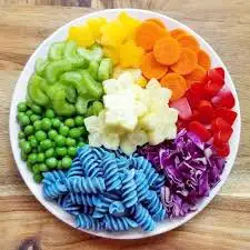 A plate of with foods each color of the rainbow. Blue pasta, purple cabbage, red veggies, orange carrots, yellow fruits, and green lima beans and peas. A pile of star shaped banana chunks are in the center.