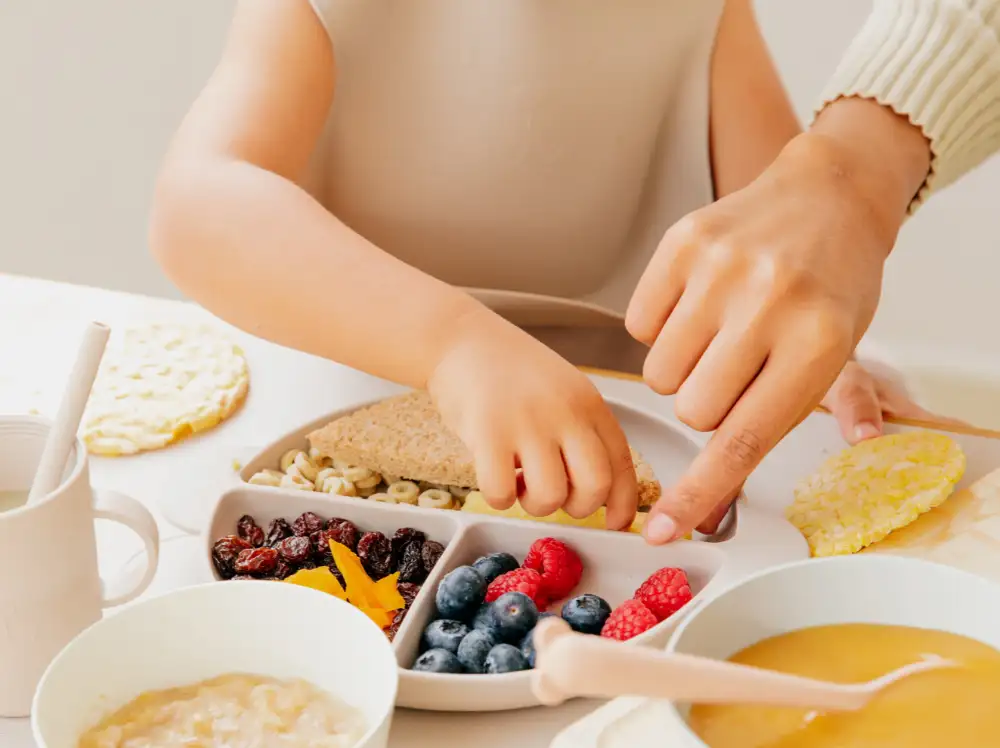 A mother and child's hands hover over a plate. The plate has sections for each group of food.